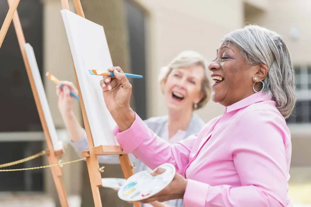 Two senior women laughing while painting at easels outdoors